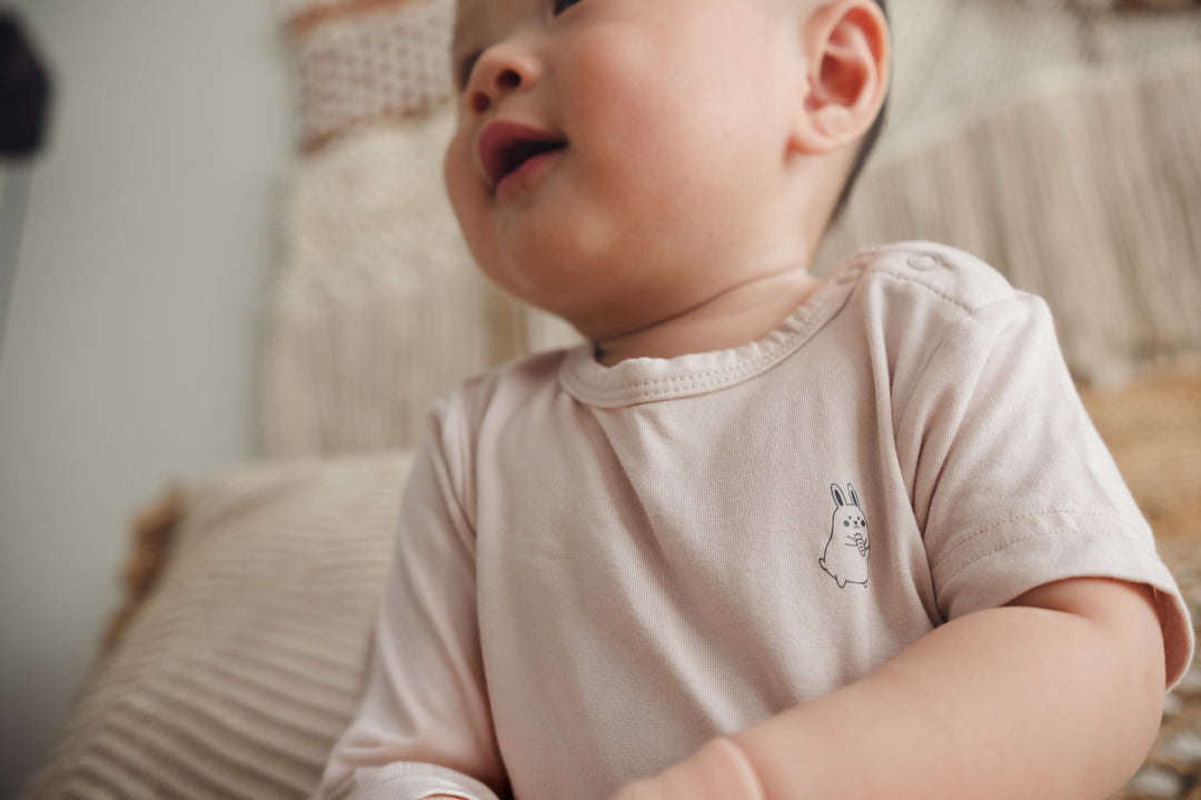 Baby sitting upright on a bed in a beige onesie