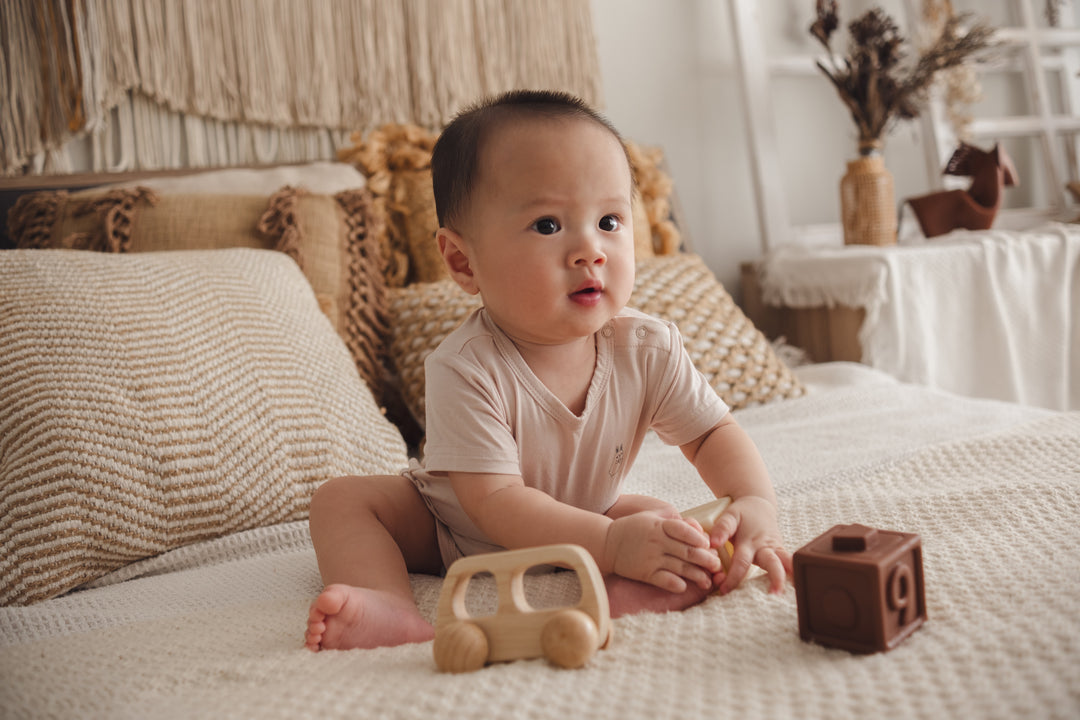 Baby in a beige onesie sitting on a bed, playing with toys in a cosy bedroom