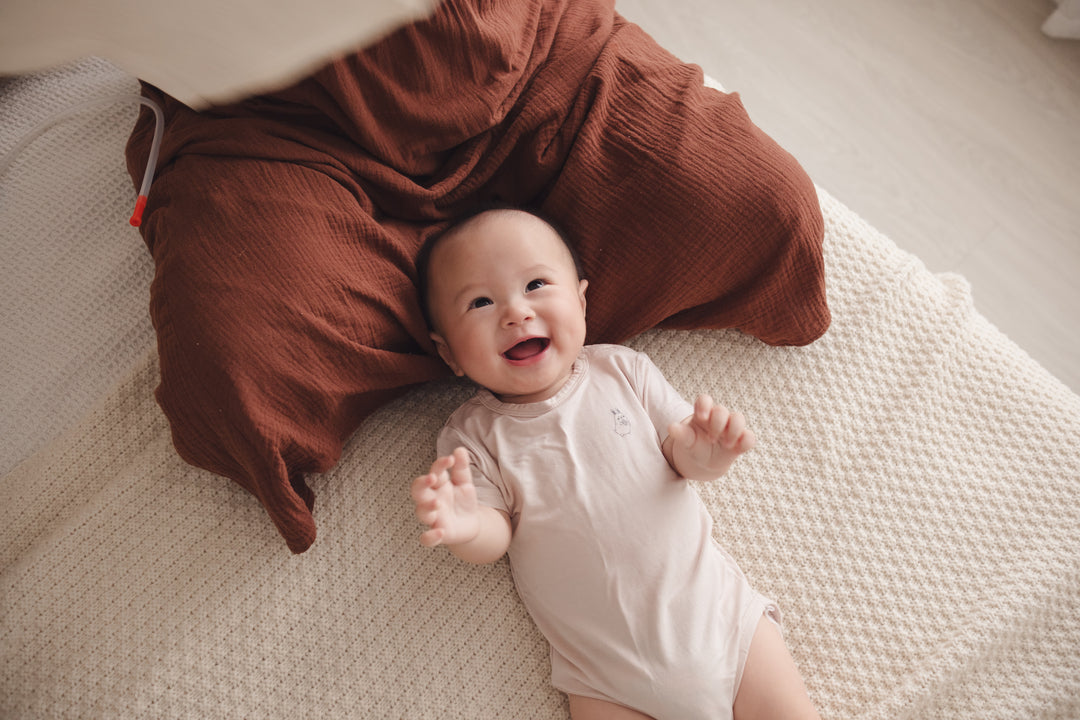 Happy baby lying on mum’s lap, wearing a beige onesie and smiling brightly