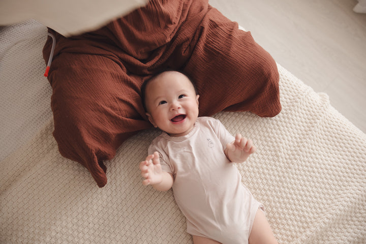 Happy baby lying on mum’s lap, wearing a beige onesie and smiling brightly