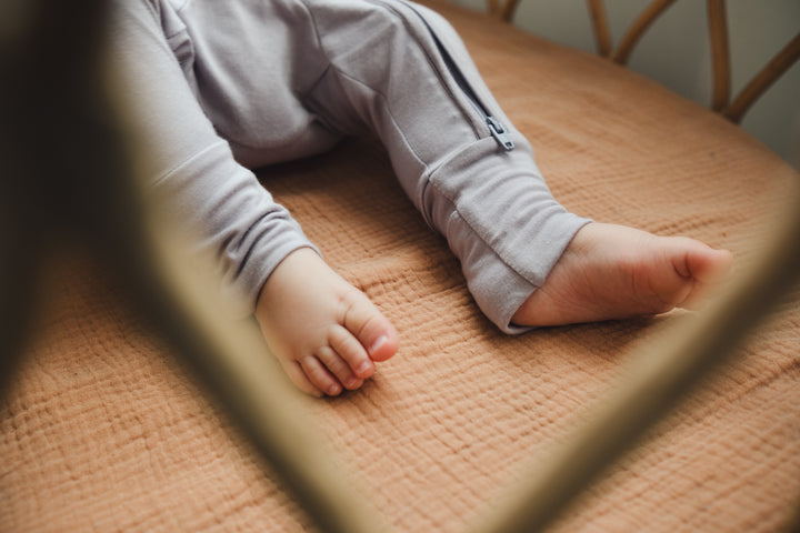 Baby lying in a crib wearing a lavender sleepsuit, showing the fold-over foot mitten detail