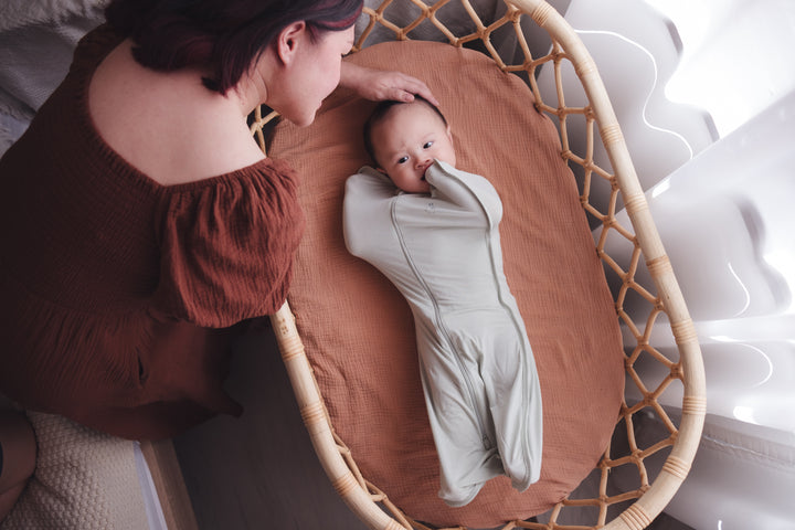 Woman leaning over a cot, gently patting the baby’s head as the baby wears a sage swaddle bag