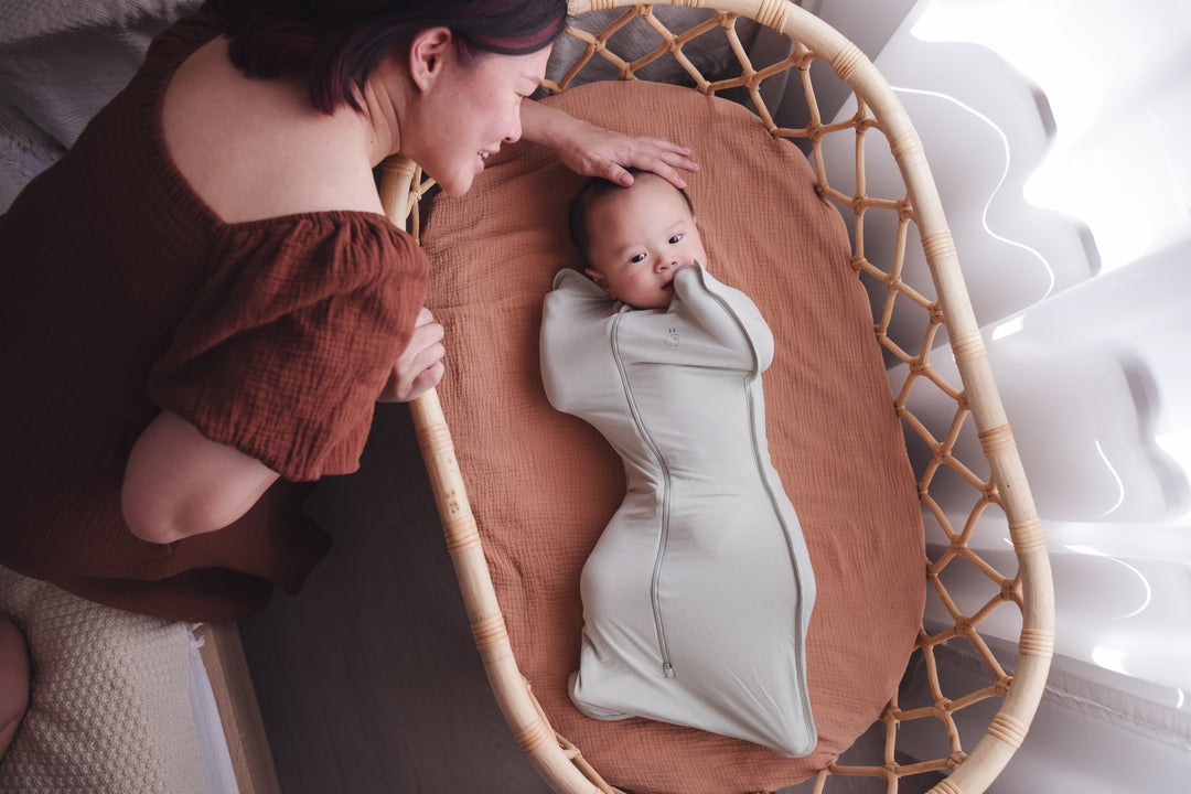 Woman leaning in to pat her baby’s head as the baby rests in a cot in a sage swaddle bag