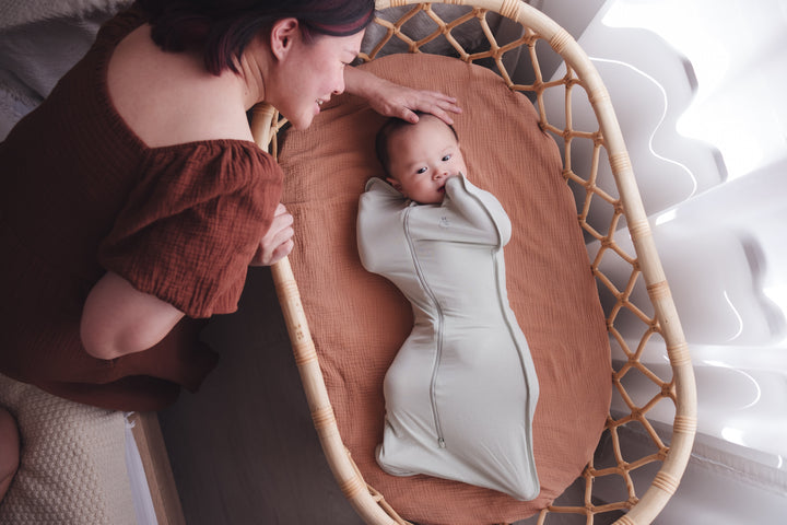 Woman leaning in to pat her baby’s head as the baby rests in a cot in a sage swaddle bag