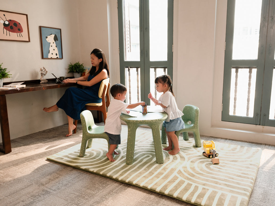 A woman sitting in a study corner while two toddlers play nearby on the Lollibly Kaya Plush Playmat.