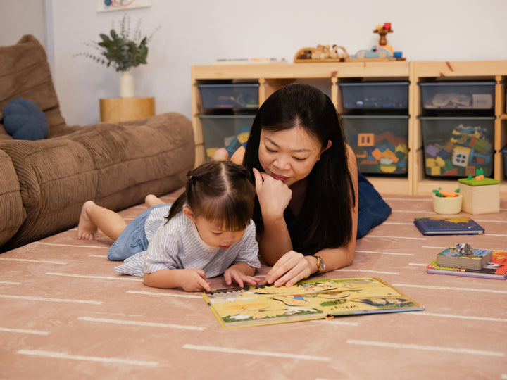 A woman and a young child lying together on the Lollibly Shortbread Plush Playmat, enjoying a colourful children’s book in a cosy living room.