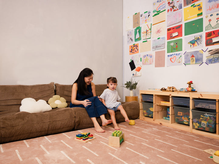 A woman and a young child sitting on a brown corduroy sofa in a cosy living room, with toys scattered across the Lollibly Shortbread Plush Playmat.