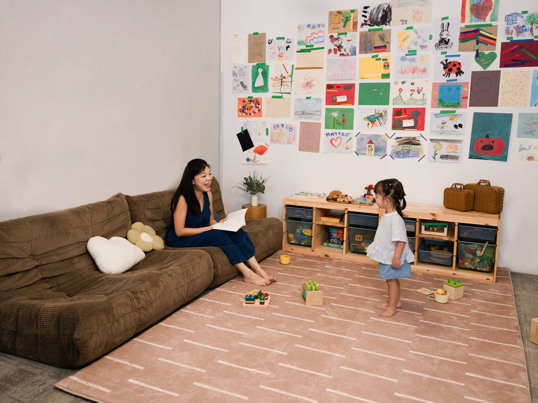 A woman sitting on a brown sofa, reading to a young child standing on the patterned Lollibly Shortbread Plush Playmat in a cosy living room.
