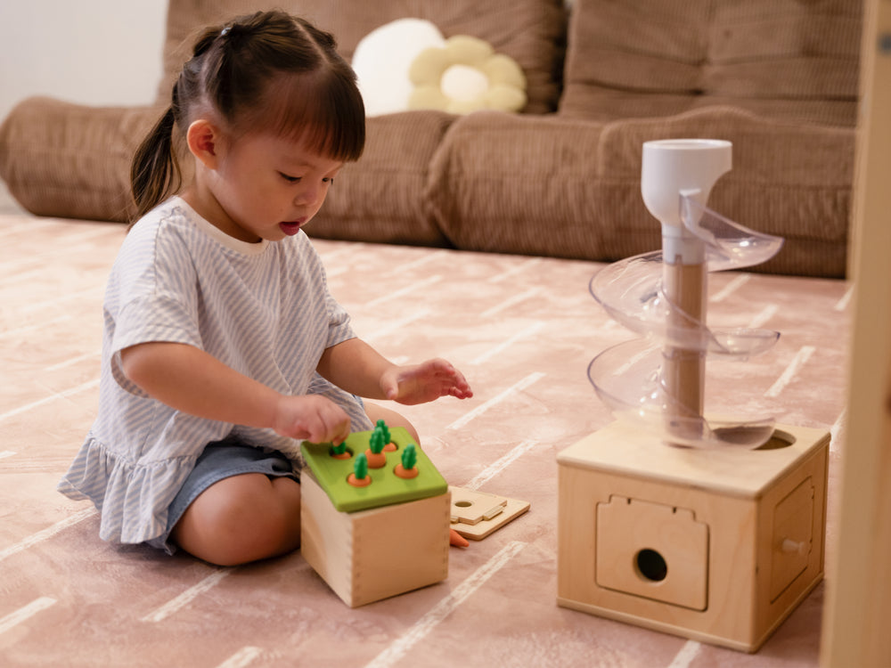 A young child sitting on the Lollibly Shortbread Plush Playmat, featuring a patterned design, while playing with wooden toys in a cosy living room.