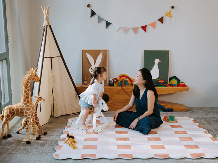 A woman and a young girl sitting together on the Lollibly Waffle checkered Plush Playmat in a cosy children’s playroom.