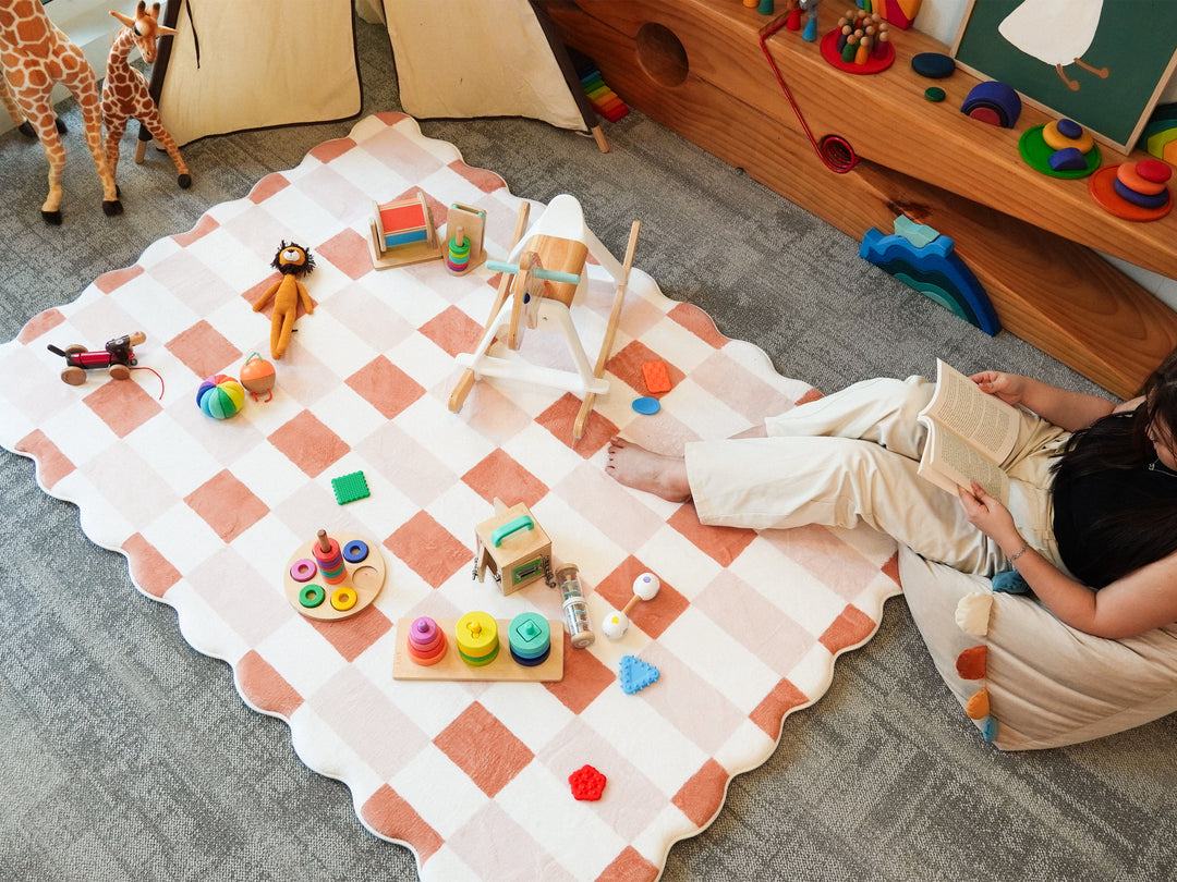 A person sitting among scattered toys on the Lollibly Waffle Plush Playmat, featuring a checkered pattern and scalloped edges in a cosy playroom.