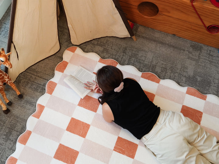 A person lying on their stomach while reading a book on the Lollibly Waffle Plush Playmat, featuring a two-tone checkered pattern and scalloped edges.